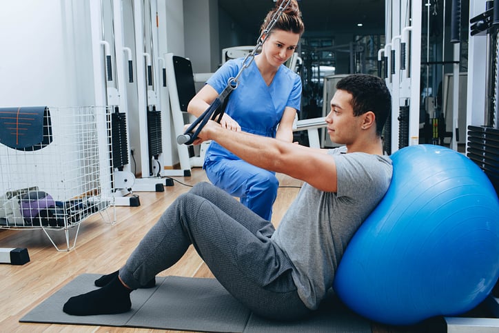 physcial therapist working with patient on cable pulldown bar with back supported by medicine ball